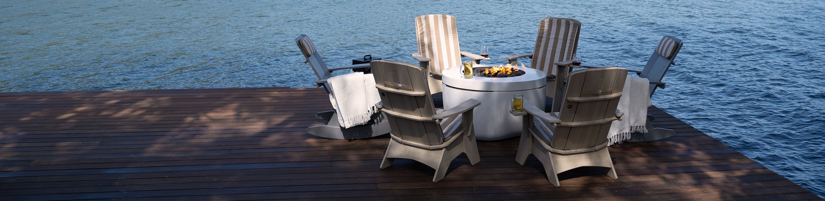 Four wooden Adirondack chairs with blankets surround a lit fire pit on a wooden deck overlooking a calm body of water. A couple of drinks are placed on the edge of the table. Shadows of trees are visible on the deck.