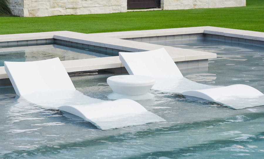 Two white Ledge Lounger Outdoor Oasis In-Pool Chaise Loungers sit partially submerged in a shallow pool with a round, white side table between them. The pools raised edge and the grassy lawn enhance this ultimate poolside essentials bundle.
