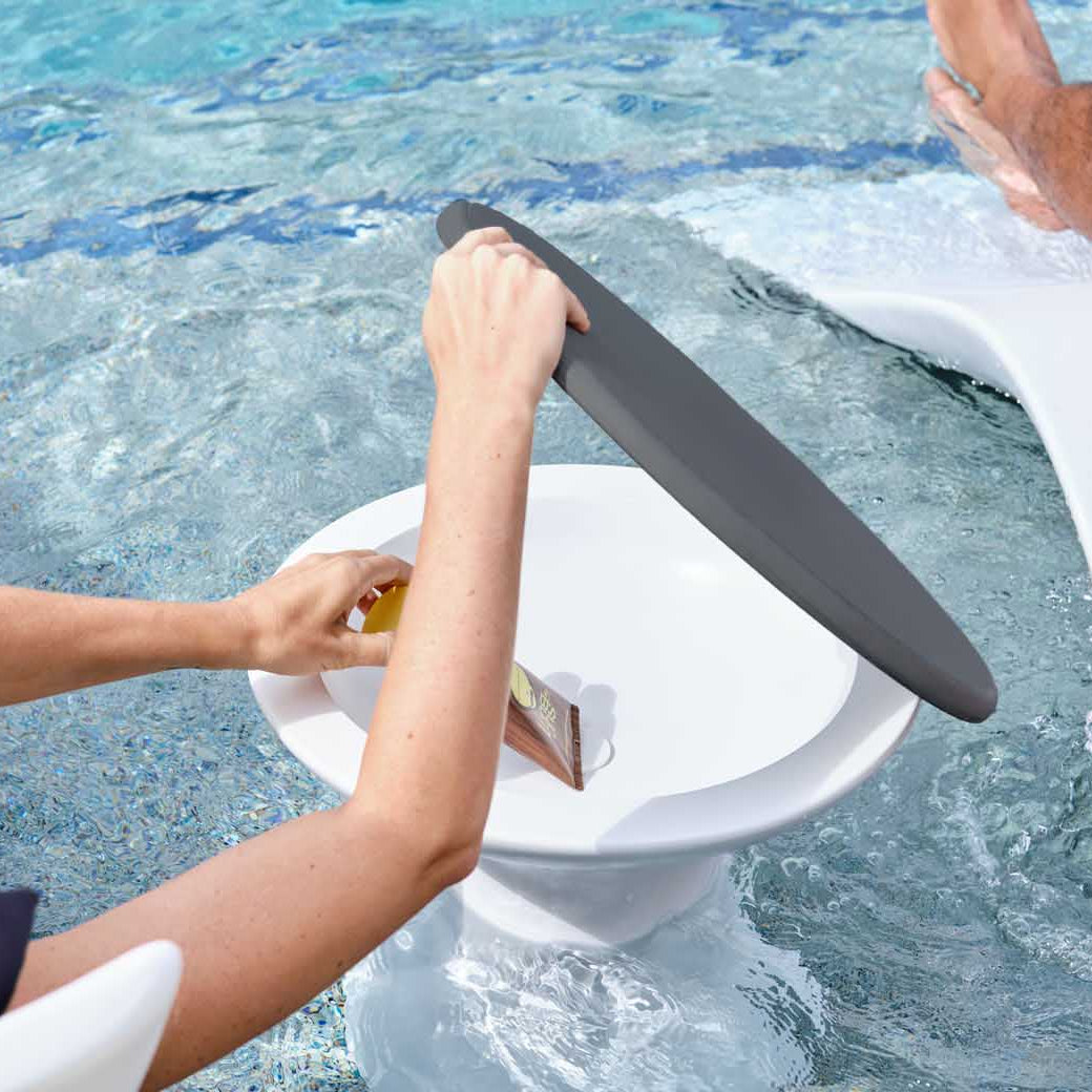 A person adjusts a black, round shade over their drink on a floating tray while enjoying the Resort Luxury Outdoor Paradise In-Pool Furniture Bundle by Ledge Lounger. Another relaxes on a chaise lounger in the water, embracing poolside leisure and sunshine.