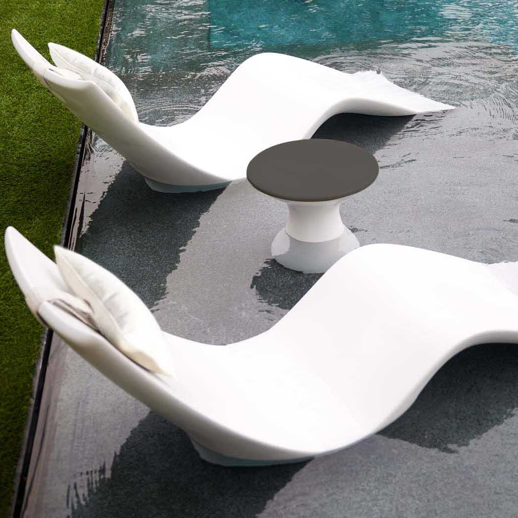 The Resort Luxury Outdoor Paradise In-Pool Furniture Bundle from Ledge Lounger features two wave-shaped, modern white chaise loungers partially submerged in a pool beside a small, black-topped round table, creating a serene and sunny poolside retreat where the pool meets the grassy lawn.