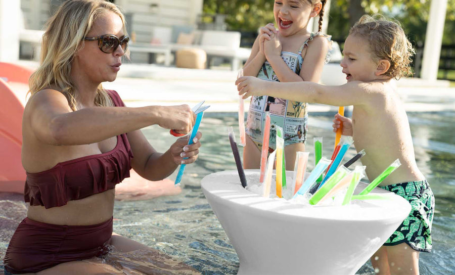 A woman in a maroon swimsuit relaxes on the Ledge Lounger Relaxation In-Pool Chair & Ice Bin Bundle, handing colorful ice pops to two nearby children. One child in a patterned swimsuit eagerly grabs an ice pop while the other smiles beside a white table with more treats under the sunny sky.