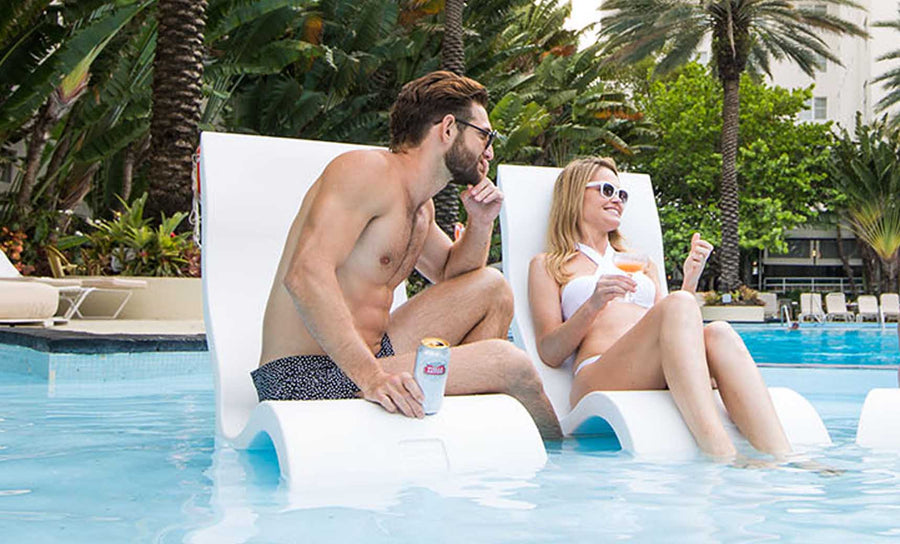 Man and woman relaxing on pool loungers partially submerged in water. The man, wearing sunglasses and swim trunks, holds a beverage can. The woman, in a white swimsuit and sunglasses, holds a drink. Palm trees and greenery are in the background.