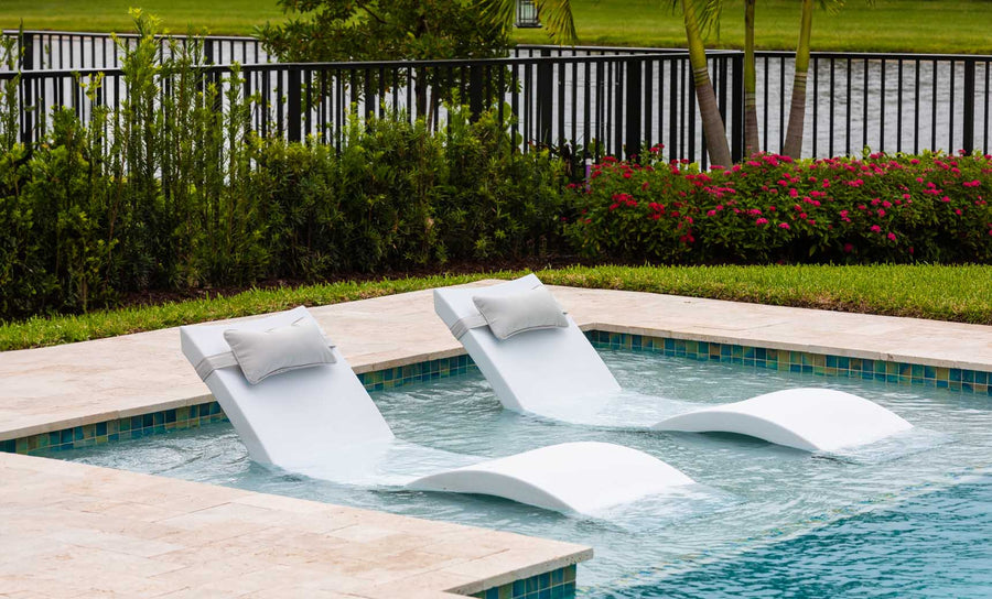 Two white chaise lounges from the Ledge Loungers Outdoor Escape In-Pool Furniture Bundle sit partly submerged in a shallow pool, facing a metal fence adorned with lush greenery and red flowers. A serene body of water and trees extend into the horizon under a clear sky.