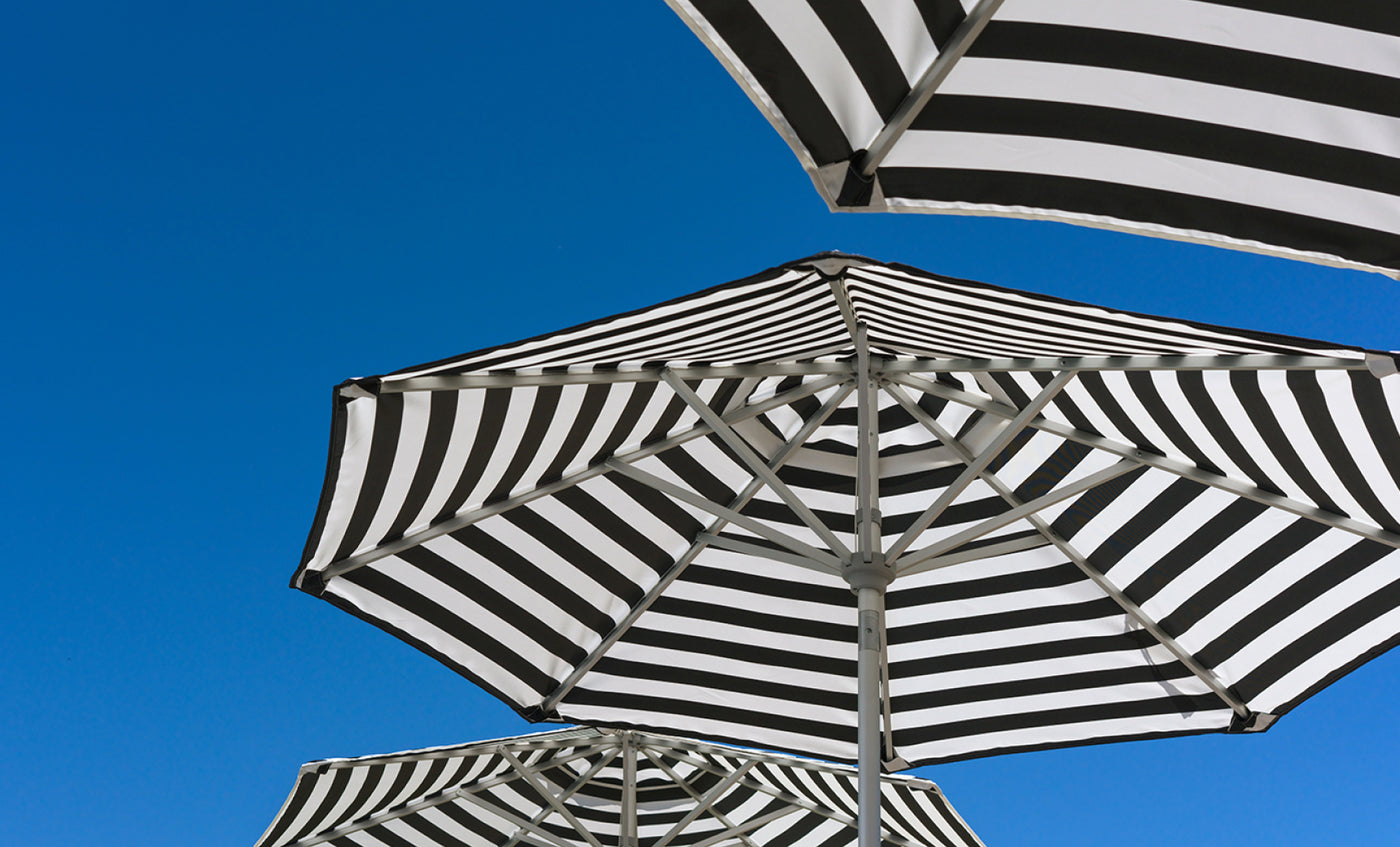 Ledge Lounger’s Signature Chaise and Side Table with Umbrella In-Pool Set features bold black and white stripes—striking geometric canopies set against a bright blue sky, perfect for enhancing your outdoor escape.