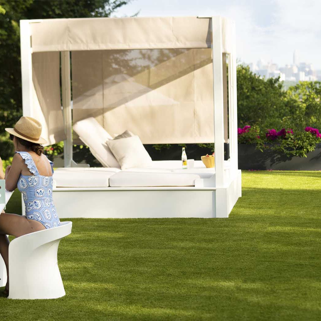 Two people sit at a white outdoor table on the grassy lawn, with a Ledge Lounger Shift Daybed nearby. One wears a sun hat and blue patterned swimsuit. Trees frame the distant city skyline, blending nature with style using durable outdoor materials.