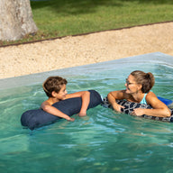 A woman and boy in the backyard pool floating on blue pool noodles.