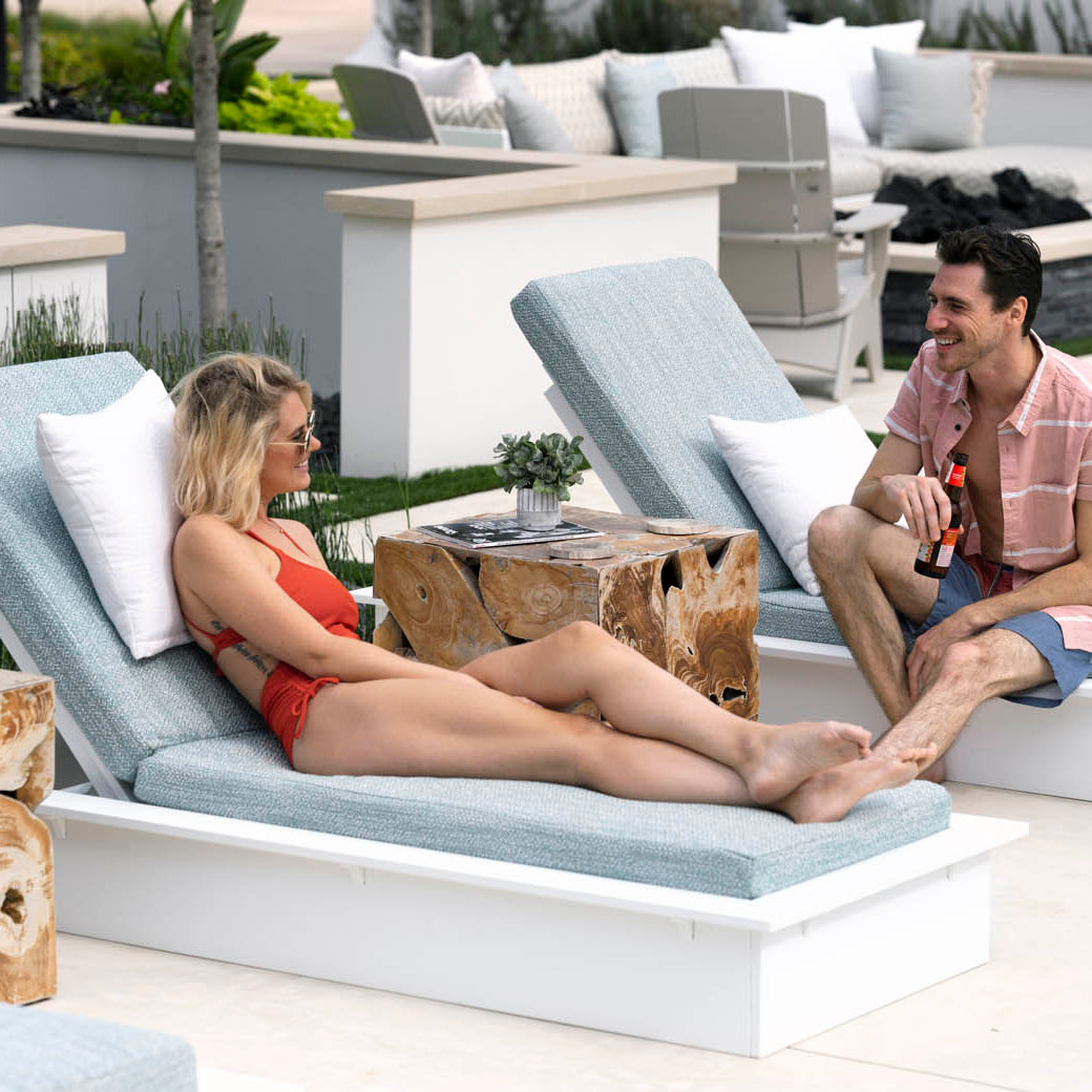 Reclining on the Ledge Lounger Echo Chaise, a woman in a red swimsuit and sunglasses smiles at a man in a pink shirt nearby. They relax among potted plants, making the most of their modern outdoor setting.