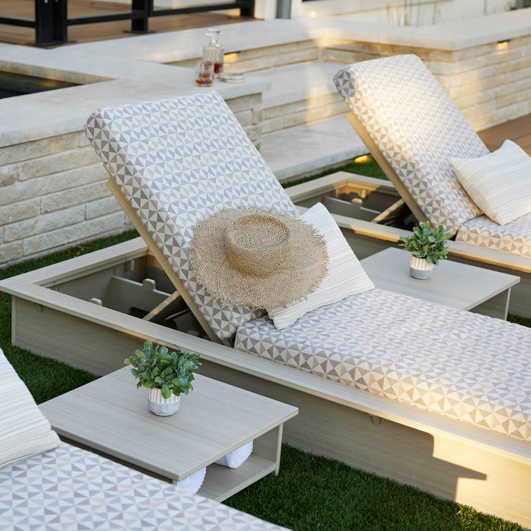 Three Echo Chaise loungers by Ledge Lounger, each with patterned cushions and a straw hat, are arranged poolside. Every adjustable chair includes a small side table with a potted plant. The area is beautifully surrounded by light stone and wooden decking.