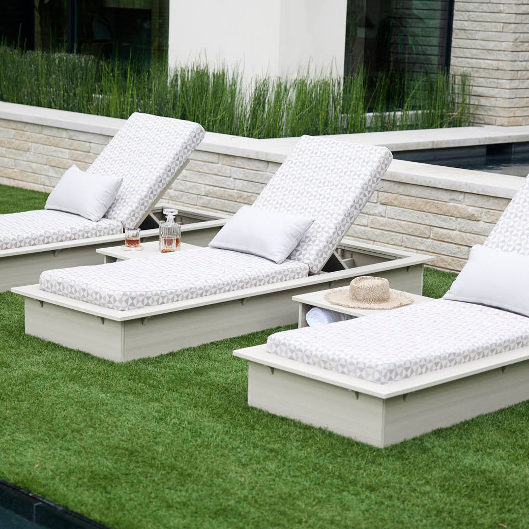 Three Echo Chaise loungers by Ledge Lounger, cushioned in white with pillows, are set on trimmed grass next to a stone-tiled pool. An adjustable chaise holds two bottles and a straw hat, enhancing the tranquil space amid lush greenery and a modern building.