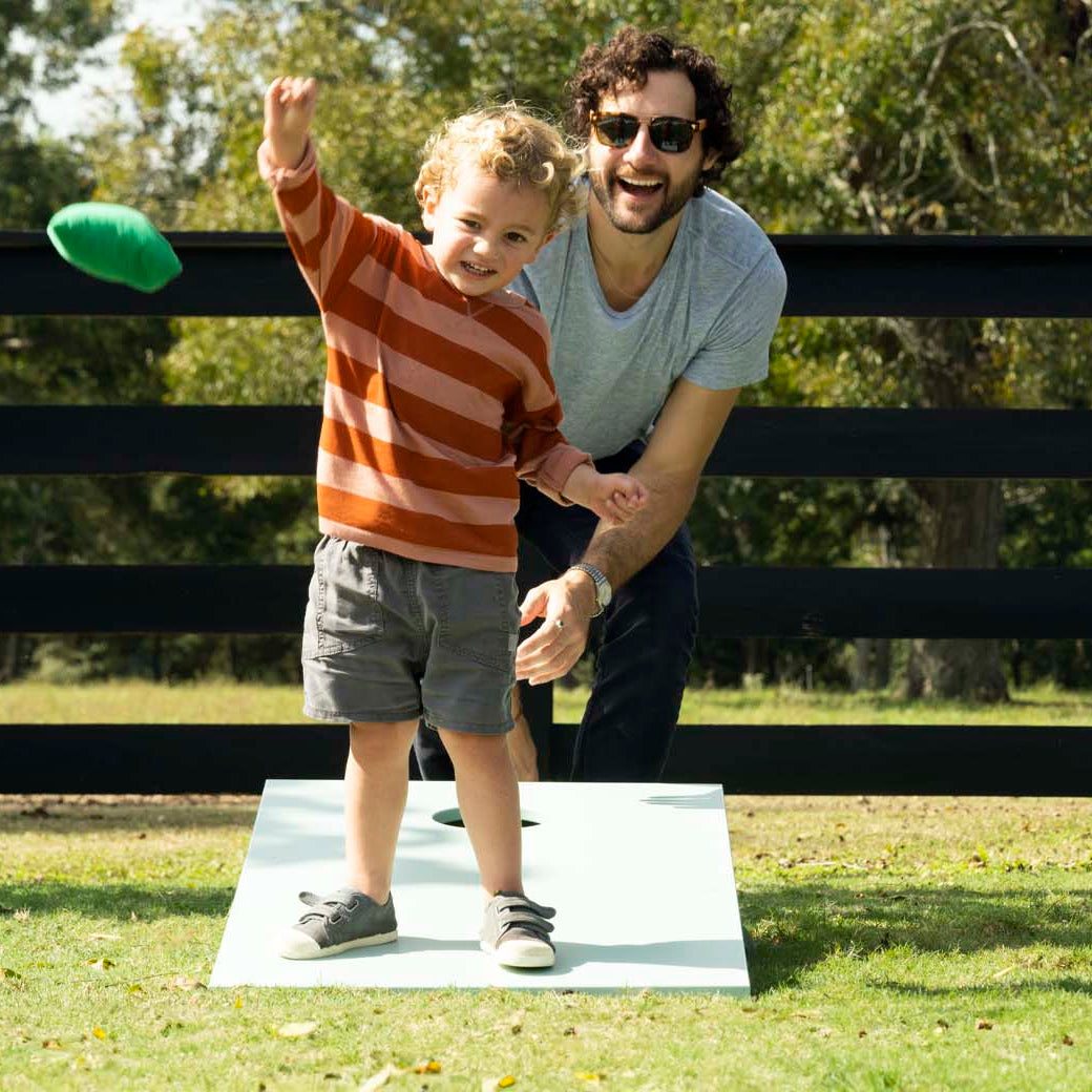 A young child wearing a striped sweater throws a bean bag while a man in sunglasses smiles and kneels beside him. They are outside on a grassy area with a black wooden fence and trees in the background.