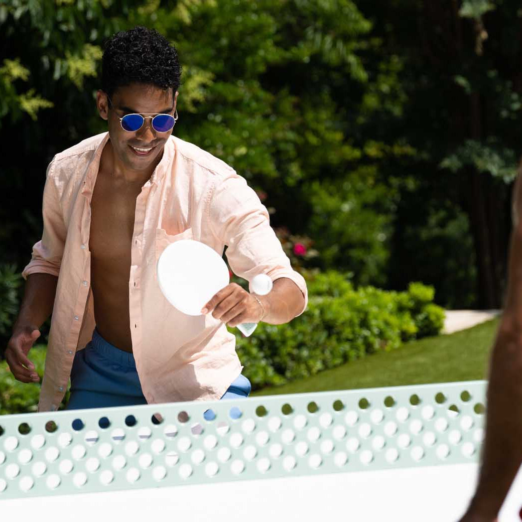 A man in a light pink shirt and sunglasses enjoys outdoor entertainment, playing ping pong with a casually dressed person. They play on a Ledge Lounger Table Tennis Set, featuring a distinct hole pattern on the net, surrounded by lush green foliage.