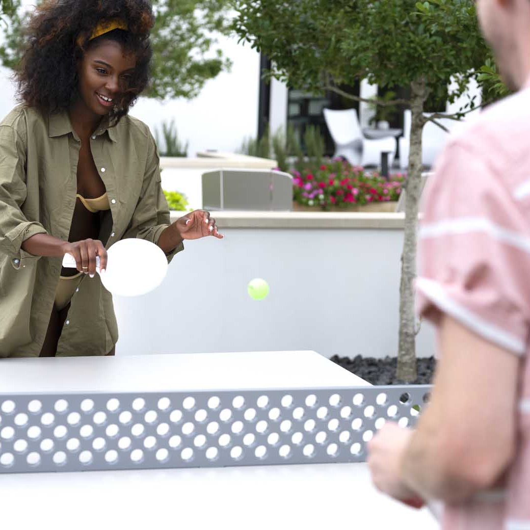 A curly-haired woman in a green jacket plays outdoor ping pong with a man in a pink shirt using the Ledge Lounger Table Tennis Set, amidst trees and flowers, ready to hit the ball. A can rests on the table nearby.