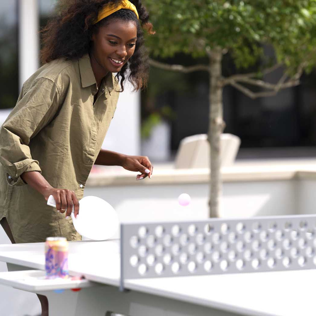 A woman in a green shirt, part of the lively Ledge Lounger Table Tennis Set, plays ping pong outdoors. Shes poised to hit the ball with precision, while a can sits nearby on the table and trees sway gently in the background.