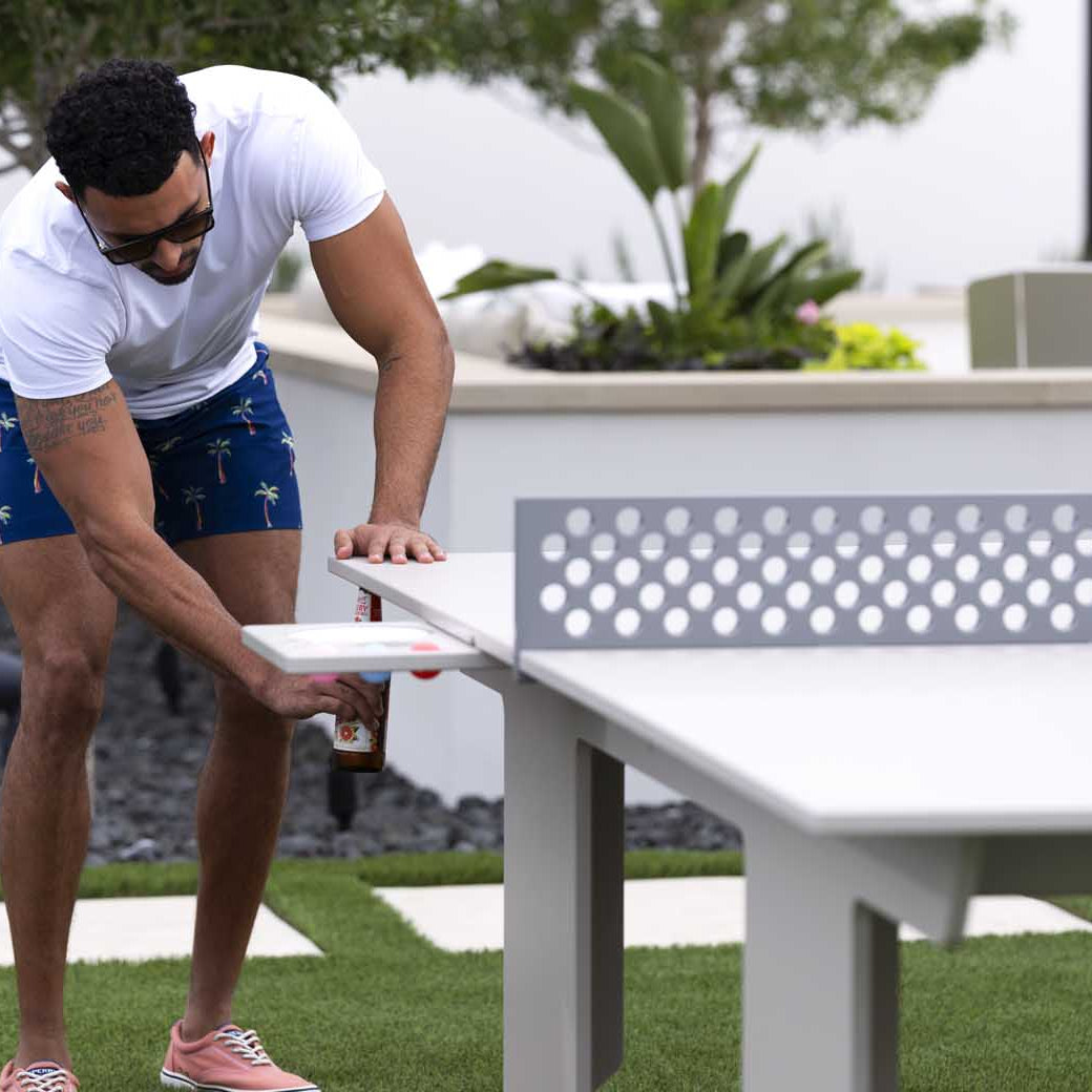A man in sunglasses and casual summer attire plays on the white Ledge Lounger Table Tennis Set. The scene unfolds in a landscaped yard filled with trees and grass, providing an ideal backdrop for outdoor entertainment.