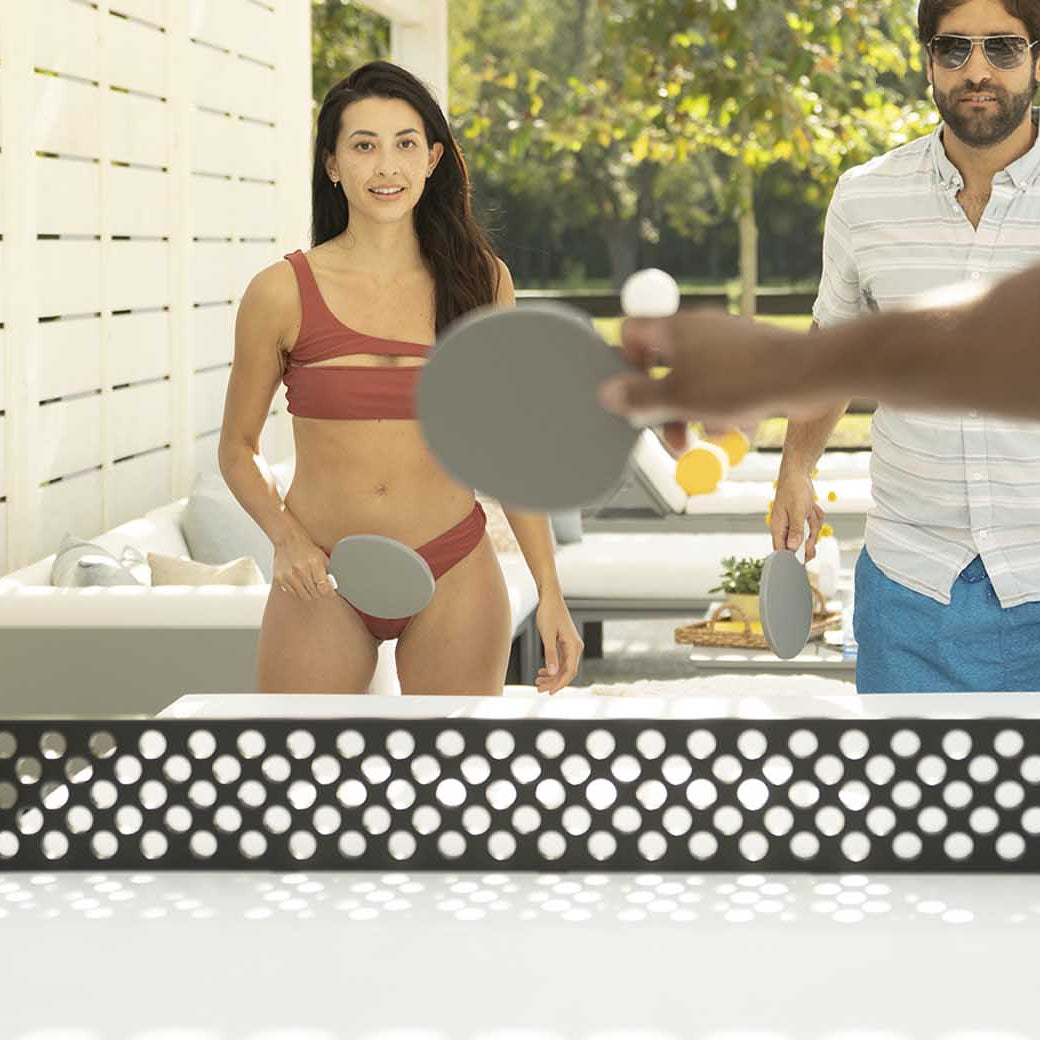 Four people enjoy outdoor entertainment, playing table tennis with the Ledge Lounger Table Tennis Set. A woman in a red bikini and a man in a striped shirt and blue shorts focus intently on the game. Trees and a white fence provide the perfect backdrop for this lively match.