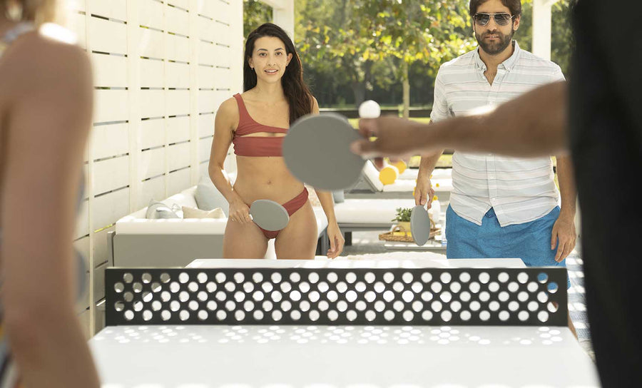 Four people enjoy outdoor entertainment, playing table tennis with the Ledge Lounger Table Tennis Set. A woman in a red bikini and a man in a striped shirt and blue shorts focus intently on the game. Trees and a white fence provide the perfect backdrop for this lively match.