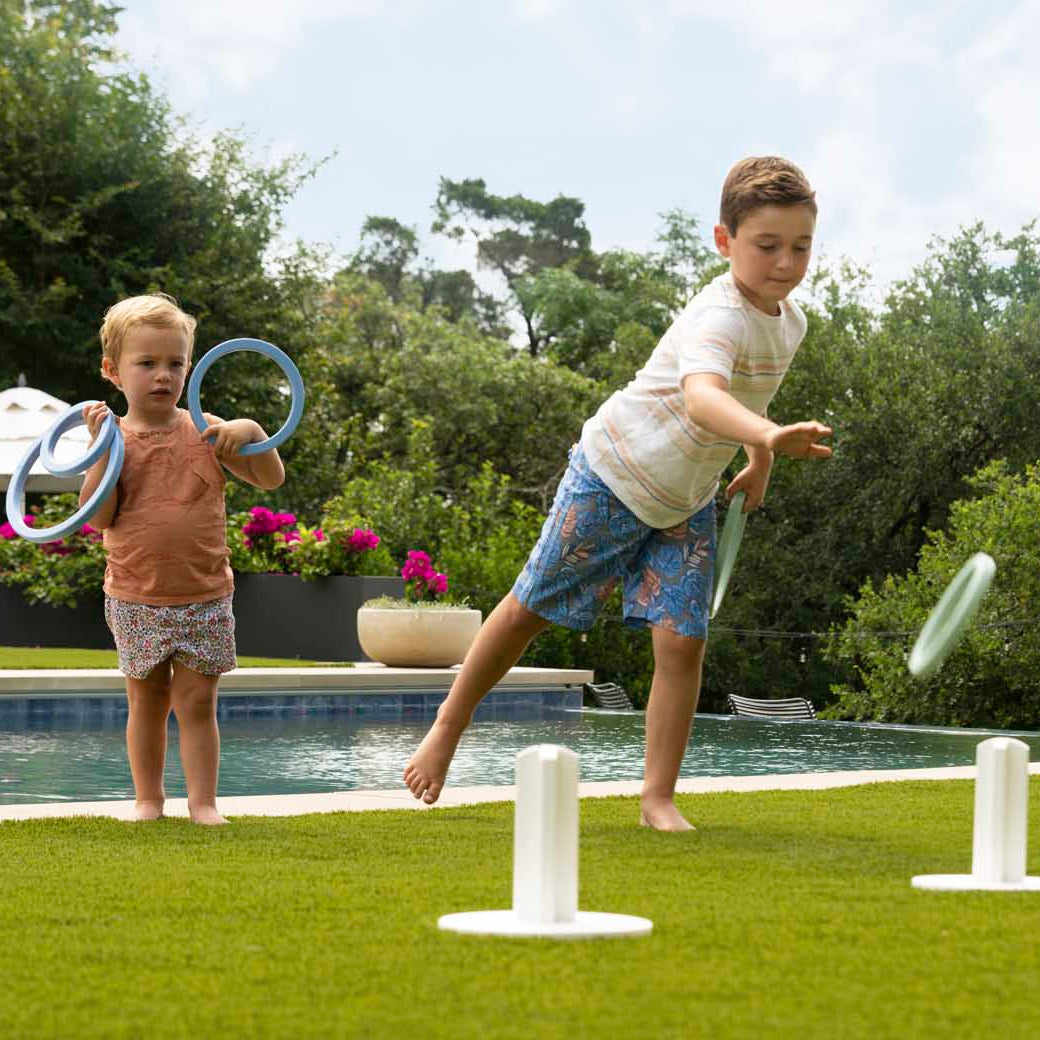 Two children play Ledge Loungers Ring Toss on a grassy lawn by a pool. The older child aims for the stakes as the younger one watches, holding rings. Trees and flowers frame this lively outdoor scene.