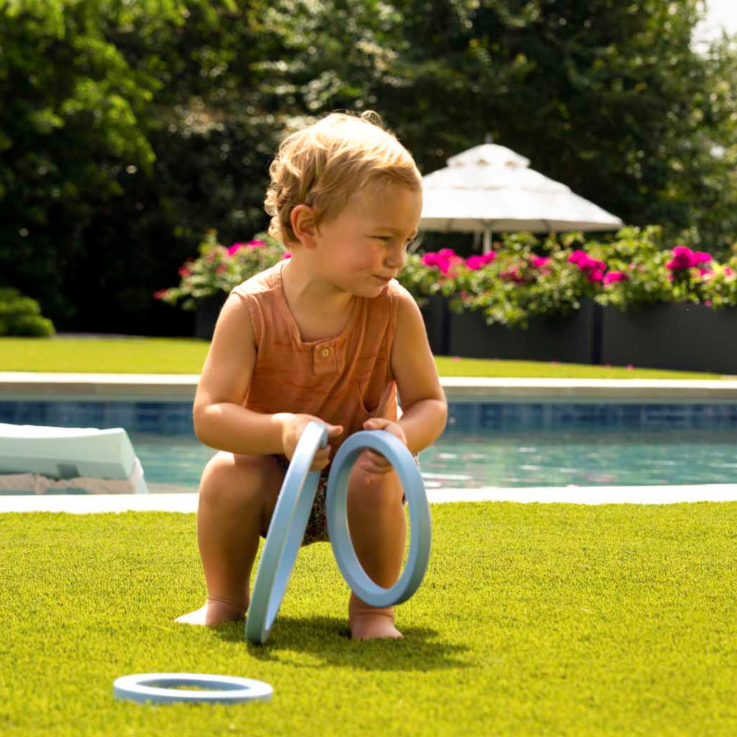 A toddler crouches on lush grass, enjoying the Ledge Lounger Ring Toss with a plastic ring by the pool. Another child balances on a circular object. Bright flowers and patio furniture add to the backyard fun in the sunny setting.