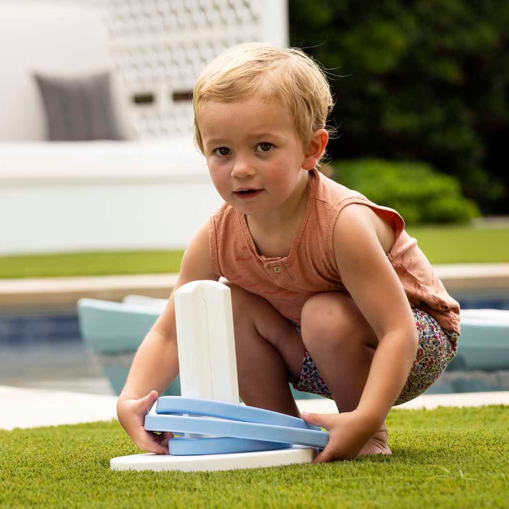 A young child with short blonde hair crouches on grass, playing Ledge Loungers Ring Toss using a blue and white plastic ring. Behind them is a pool and a cushioned seating area amidst greenery. The child sports a sleeveless orange top and patterned shorts—ideal for backyard fun.