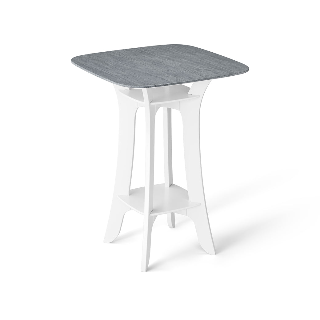 The Haven Hightop Table by Ledge Lounger features a square, light blue-grey tabletop with a white, modern base, curved legs, and a lower shelf. Its durable, minimalist design is contemporary and perfect for any patio collection.