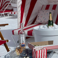 In a snowy outdoor setting, red and white striped lounge chairs create a cozy scene. A canopy shelters a table with delights: wine, champagne chilling in a Ledge Lounger Signature Ice Basin, snacks, and a glass. Pillows complete the inviting winter tableau.