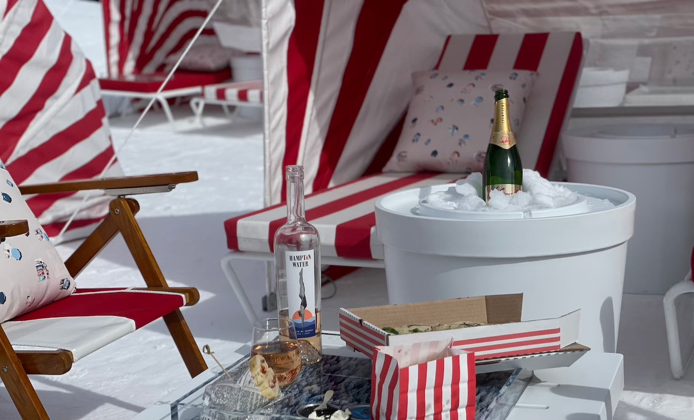 In a snowy outdoor setting, red and white striped lounge chairs create a cozy scene. A canopy shelters a table with delights: wine, champagne chilling in a Ledge Lounger Signature Ice Basin, snacks, and a glass. Pillows complete the inviting winter tableau.