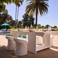 A contemporary Ledge Lounger Table Tennis and Barstools Gathering Set is arranged by a swimming pool, surrounded by palm trees, lounge chairs, and a sun umbrella under sunny skies.