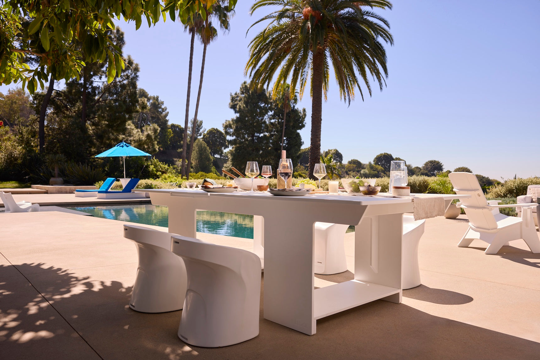 A contemporary Ledge Lounger Table Tennis and Barstools Gathering Set is arranged by a swimming pool, surrounded by palm trees, lounge chairs, and a sun umbrella under sunny skies.