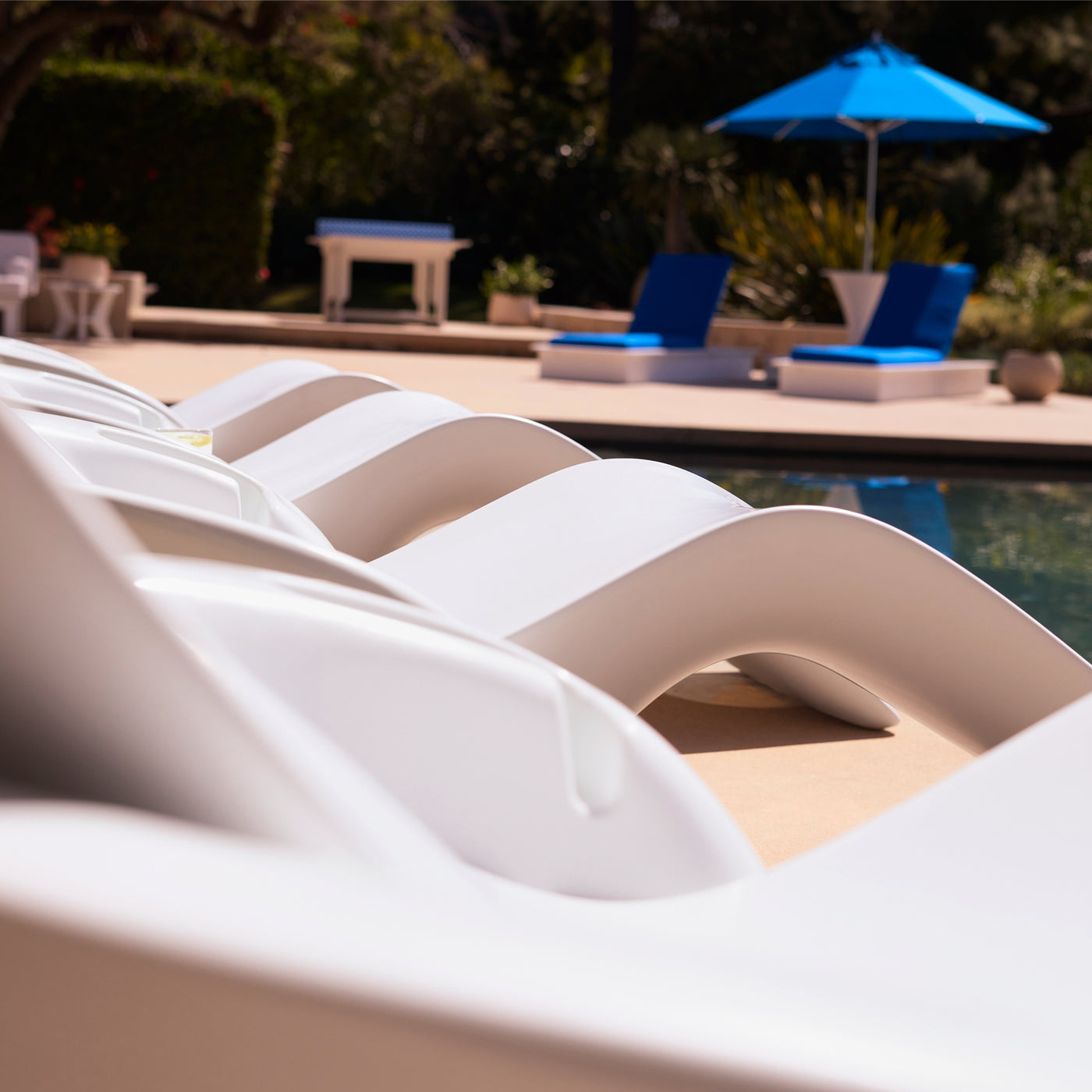Curved white lounge chairs line the edge of a pool, with blue cushioned chairs and a blue umbrella in the background, set in a sunny outdoor area with greenery.