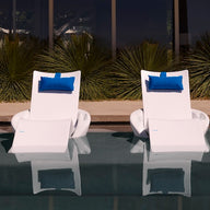 The Ledge Lounger Autograph Chaise Island Lounge Set features two modern white chaise lounges with blue pillows, partially submerged in a pool and set against large windows, surrounded by lush green plants.