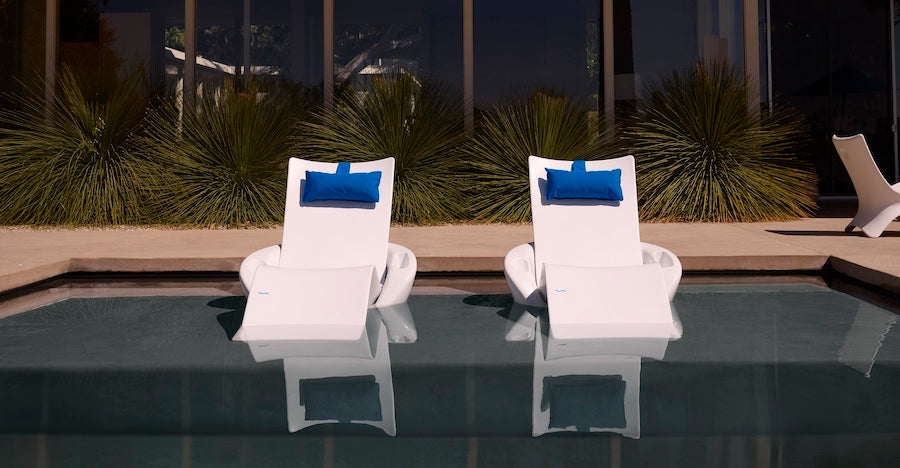 The Ledge Lounger Autograph Chaise Island Lounge Set features two modern white chaise lounges with blue pillows, partially submerged in a pool and set against large windows, surrounded by lush green plants.