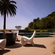 Ledge Lounger Autograph Chairs surround a round fire pit by the pool on a sunny patio, with lush greenery and a palm tree in the background under a clear sky.