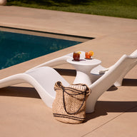 The Ledge Lounger Autograph Chaise, with lumbar support, sits poolside next to a matching table holding two drinks. A striped towel hangs on the chair and a woven bag rests nearby—ideal for pool owners seeking comfort and style.