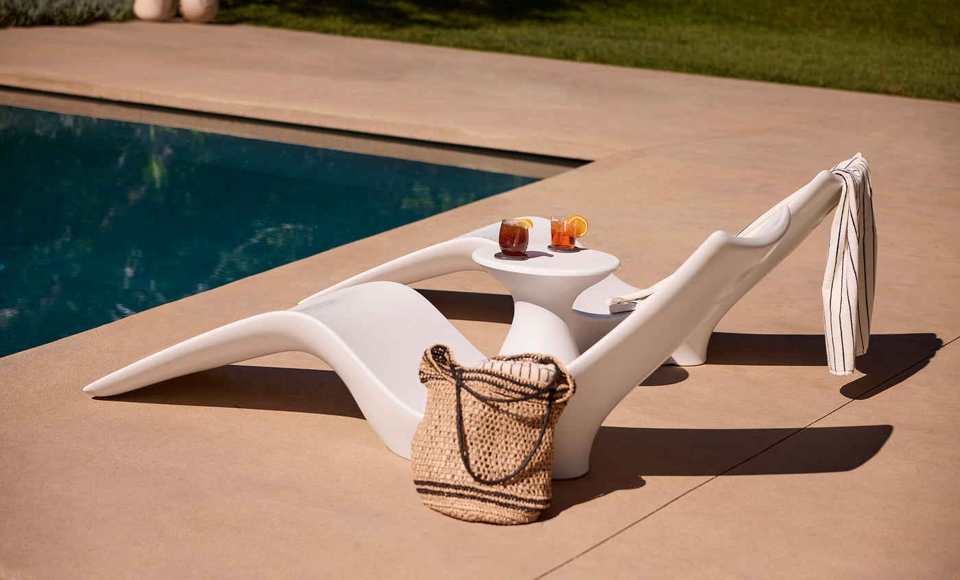 The Ledge Lounger Autograph Chaise, with lumbar support, sits poolside next to a matching table holding two drinks. A striped towel hangs on the chair and a woven bag rests nearby—ideal for pool owners seeking comfort and style.