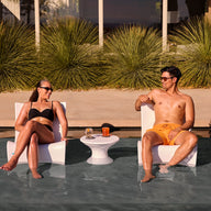 A woman and man in swimsuits relax on Ledge Lounger Autograph Chairs in a shallow pool, facing each other and smiling. A small table with drinks is between them, with tall green plants and a building in the background.