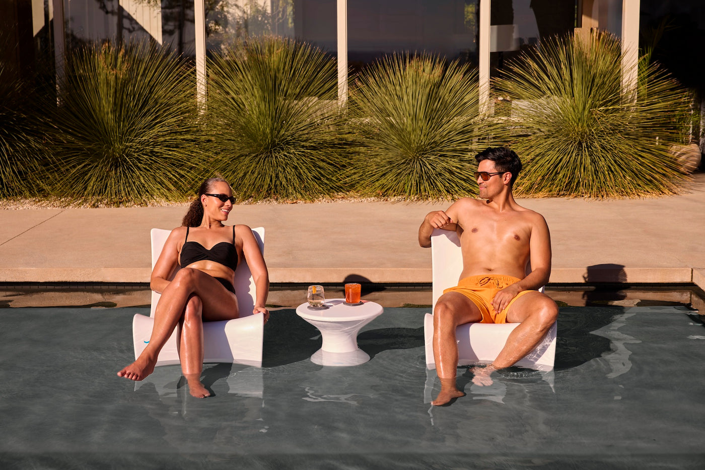 A woman and man in swimsuits relax on Ledge Lounger Autograph Chairs in a shallow pool, facing each other and smiling. A small table with drinks is between them, with tall green plants and a building in the background.