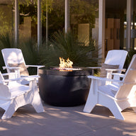 The Ledge Lounger Halo Firepit with Set of 4 Adirondacks features four white UV-rated resin chairs with all-weather resistance, arranged around a black fire pit on a patio beside a modern glass building and lush green plants.