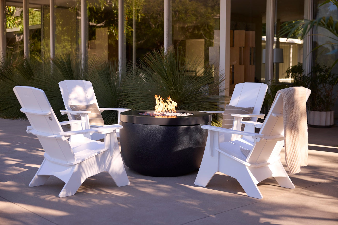 The Ledge Lounger Halo Firepit with Set of 4 Adirondacks features four white UV-rated resin chairs with all-weather resistance, arranged around a black fire pit on a patio beside a modern glass building and lush green plants.