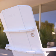 Close-up of the back of the Ledge Lounger Mainstay Ultimate Adirondack, featuring a built-in metal bottle opener, set on a patio in front of a modern building with large windows and lush greenery.