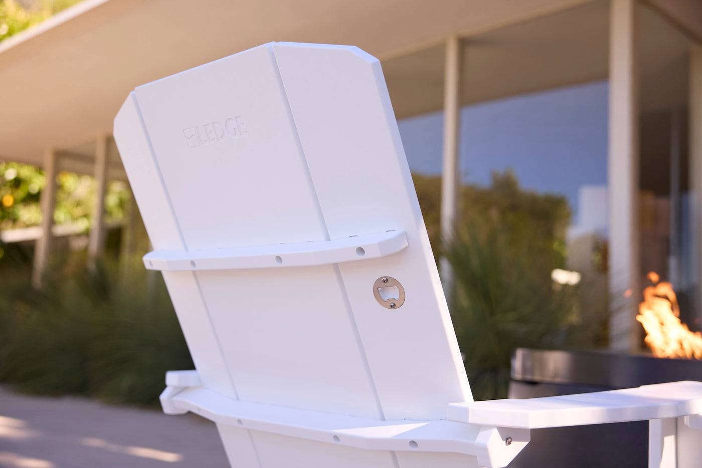 Close-up of the back of the Ledge Lounger Mainstay Ultimate Adirondack, featuring a built-in metal bottle opener, set on a patio in front of a modern building with large windows and lush greenery.