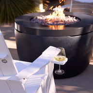 A glass of white wine sits on the armrest of a Mainstay Ultimate Adirondack by Ledge Lounger beside a modern black fire pit with flames and stones, surrounded by more white outdoor chairs on a stylish patio.