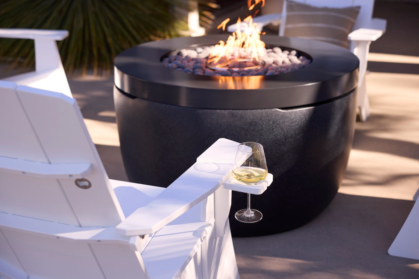 A glass of white wine sits on the armrest of a Mainstay Ultimate Adirondack by Ledge Lounger beside a modern black fire pit with flames and stones, surrounded by more white outdoor chairs on a stylish patio.