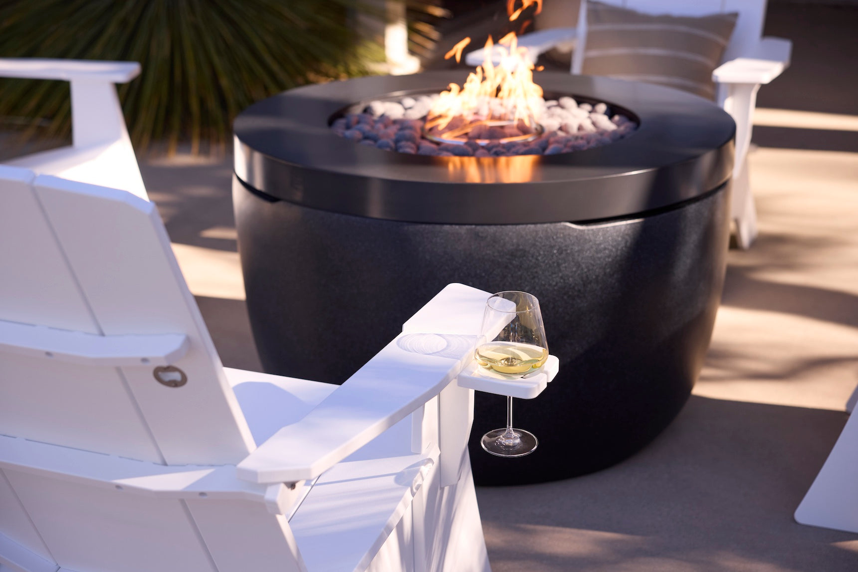 A glass of white wine sits on the armrest of a Mainstay Ultimate Adirondack by Ledge Lounger beside a modern black fire pit with flames and stones, surrounded by more white outdoor chairs on a stylish patio.