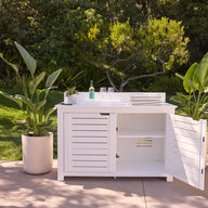 The Ledge Lounger Mainstay Bar Credenza, shown with one door open, stands on a patio between potted plants. Its countertop displays a wine bottle, glasses, and towels, set against lush greenery and trees in the background.