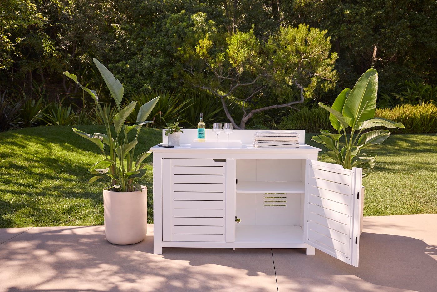 The Ledge Lounger Mainstay Bar Credenza, shown with one door open, stands on a patio between potted plants. Its countertop displays a wine bottle, glasses, and towels, set against lush greenery and trees in the background.