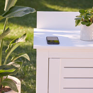 The Mainstay Bar Credenza by Ledge Lounger, styled as a white outdoor table, displays a potted plant, smartphone, and bottle. Nearby, a large green plant and grassy lawn enhance the cozy patio ambiance.
