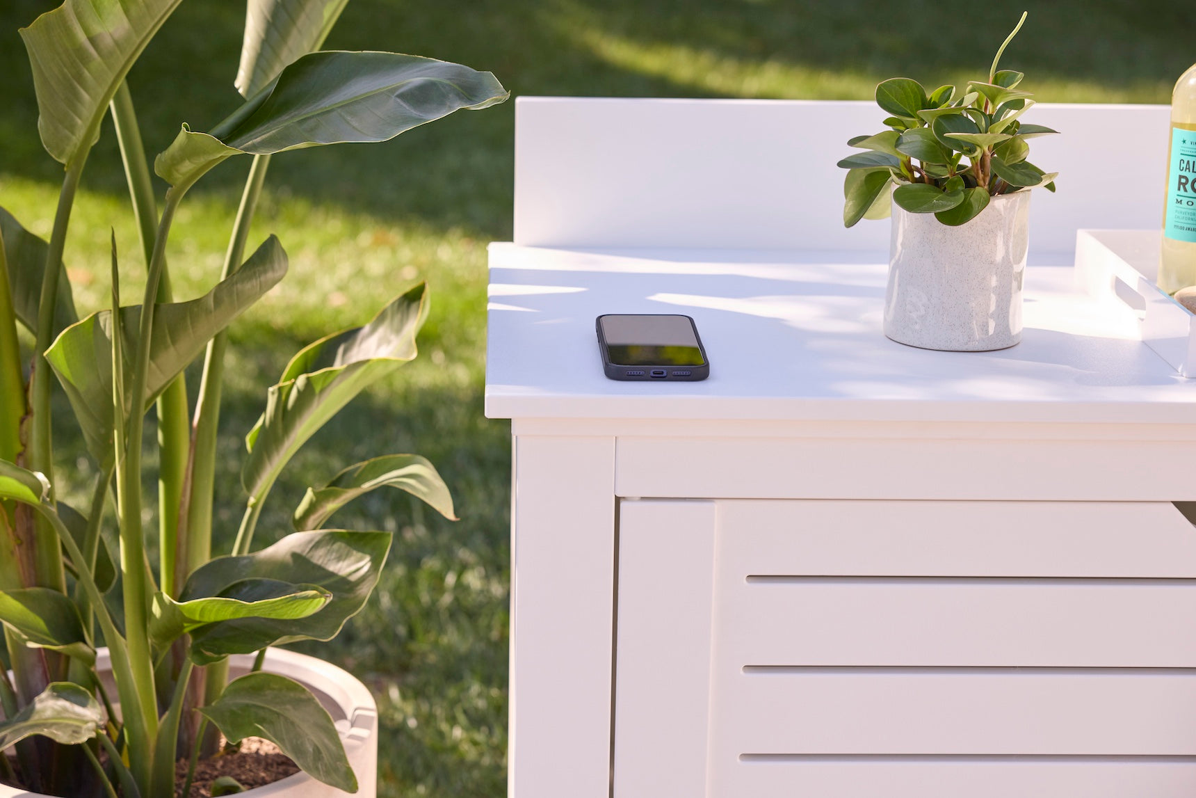The Mainstay Bar Credenza by Ledge Lounger, styled as a white outdoor table, displays a potted plant, smartphone, and bottle. Nearby, a large green plant and grassy lawn enhance the cozy patio ambiance.