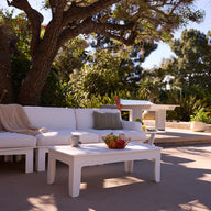 The Ledge Lounger Mainstay Sectional 3 Piece Sofa, with green pillows and a beige throw, sits under a large tree beside a white coffee table holding fruit and a cup, surrounded by lush greenery on the sunny patio.
