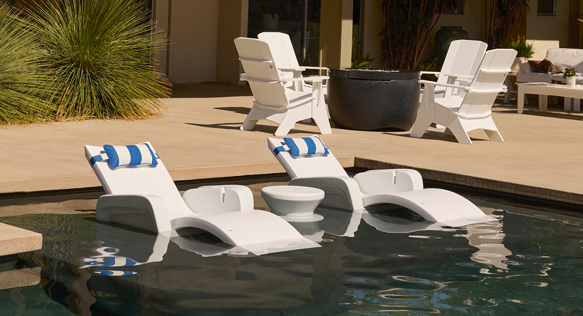 White outdoor lounge chairs with blue striped pillows arranged around a swimming pool, with patio seating and plants in the background.