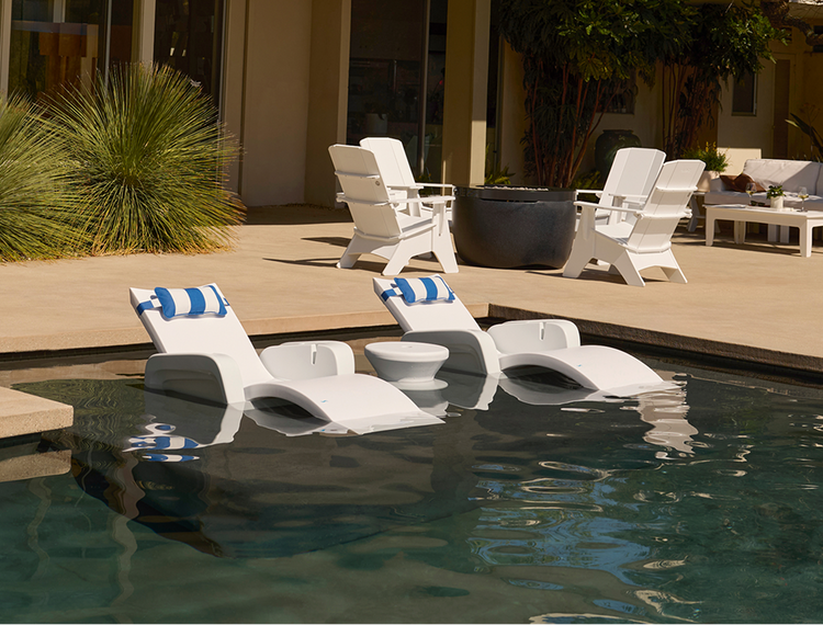 White outdoor pool lounge chairs with blue striped pillows arranged around a swimming pool on a patio, with a modern house and plants in the background.